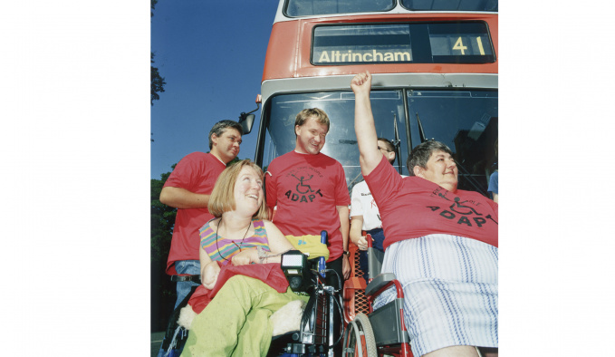 Five people, 2 are wheelchair users, are in front of a number 41 bus to Altrincham. They are smiling and looking to their left. Some of them have red t-shirts on with the word ADAPT under a wheelchair logo. 