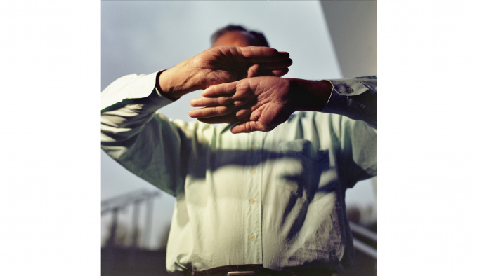 A man is photographed from the waist up. He is holding his hands up and out, with palms facing the viewer. His hands obscure his face. He is wearing a white/cream shirt and behind him are grey clouds.