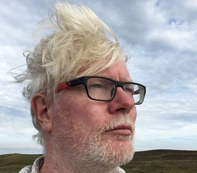 Aidan Moesby wearing dark rimmed glasses looks to the right. His hair is wind swept. He stands outside against a blue but cloudy sky