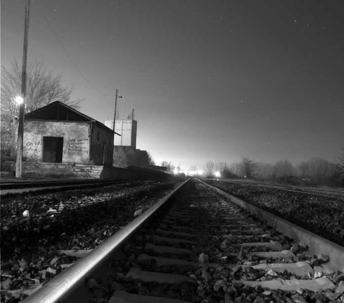 An empty railway track at night. The tracks lead the viewer from the centre bottom on the image into the distance. On the left hand side are buildings that look deserted.