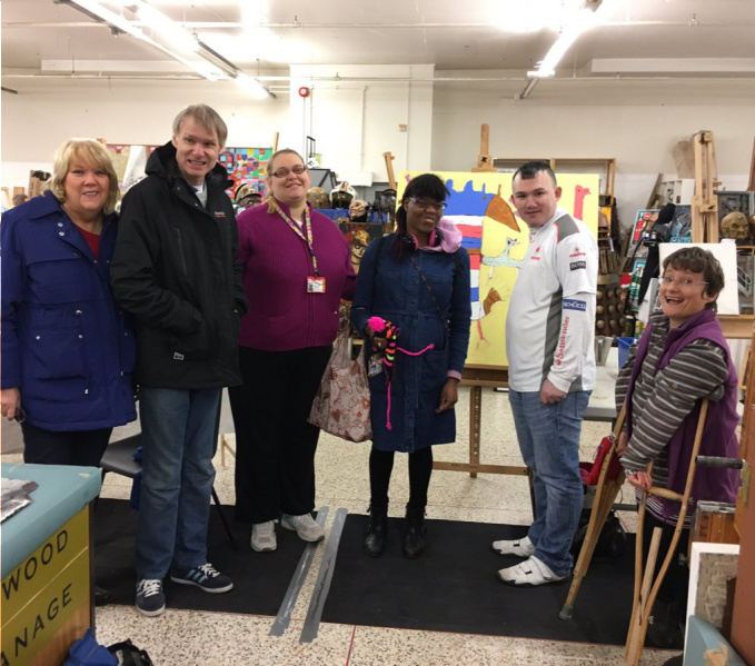Six of the ArtStudio01 artists in their studio. They are all looking at the camera and smiling, behind them are easels with artworks in progress.