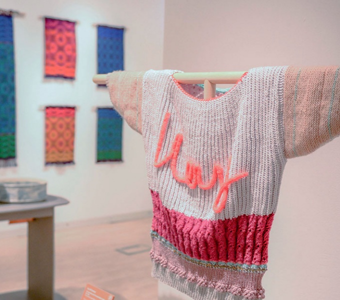 A pink woollen hand knitted jumper is displayed in a gallery space. On the wall behind is an assortment of woven art works.
