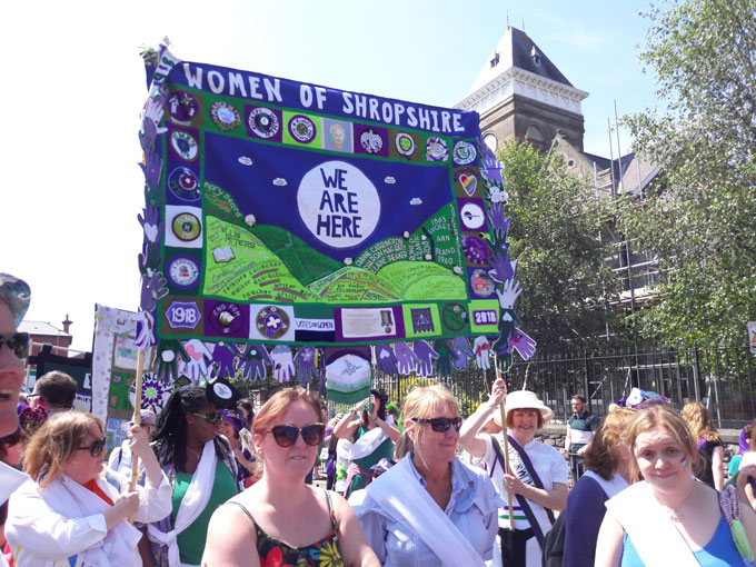 Images shows a close up of a larger crowd of women carrying a textile banner, made from purple, white and green fabrics. The main text of the banner reads 'Women of Shropshire' horizontally across the top. Beneath it reads We Are here in a circle. Below that are the shapes of hills with the names of women of noteembroidered on to them.