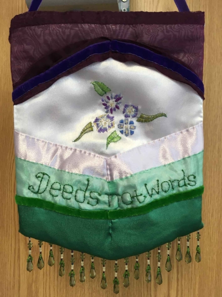 Embroidery pennant created by Rose Foran for the DASH PROCESSIONS project.  Using the colours of the suffrage movement, violet, white and green, the pennant is horizontally striped, using silk, satin and ribbon, it has flowers and 'Deeds not words' embroidered into the fabric. Delicate green beads finish the pennant at the bottom edge. 
