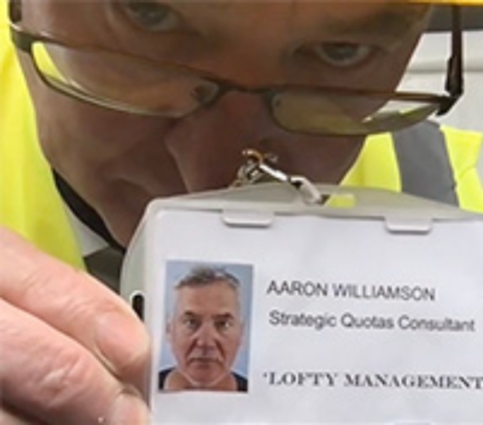 A man in glasses and a yellow builders helmet leans towards the camera holing up a name badge. It reads Aaron Williamson, Strategic Quotas Consultant, Lofty Management