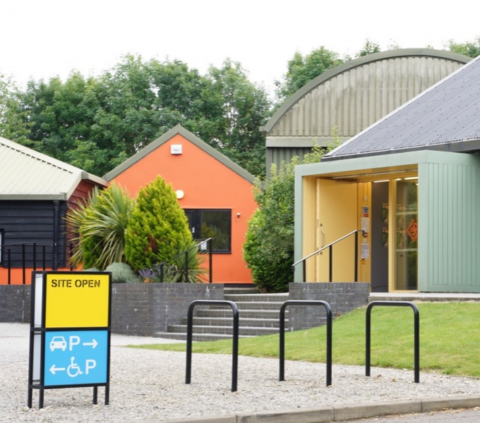 Exterior shot of Wysing Arts Centre. Foreground - bicycle racks and 'Open' sign, behind this are steps leading to orange, green and grey buildings, trees frame the buildings in the background.