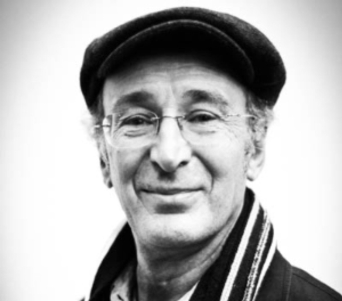 A black and white portrait photograph of Mike Layward. He wears a flat cap, a stripey scarf and frameless glasses as he faces the camera.