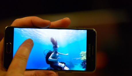 A left hand is holding a mobile on which Sue Austin is seen underwater swimming in her wheelchair with scuba tanks on her back. the sea around her is beautiful cyan blue with her dark ponytail hair flowing up into reflected sunlight.