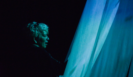 Against a black background, a womans side on face is lit with blue cyan light with the pieces of stretched fabric also lit with projected light, in front of her face, on the right of the photo.