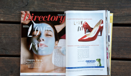 A centre spread of a women's magazine on a wooden bench. On the left hand page is a woman with a face mask on. The words 'Love Me' have been cut out of her face. On the right hand page there is a pair of red high heeled court shoes.