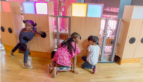 three young children with black hair are listening , crouched with ears to the black, round ear speakers on the wooden panels, with square lit squares in yellow, cyan, pink and blue at the top of the wooden panels.