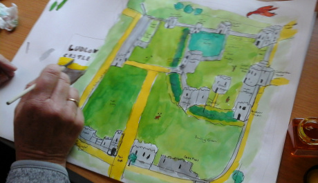 A white paper, centrally covered in a green watercolour showing a map being created of Ludlow Castle grey buildings, with a yellow boundary pathways ,  with a hand holding a pantbrush in a right handwriting the words Ludlow Castle in grey.