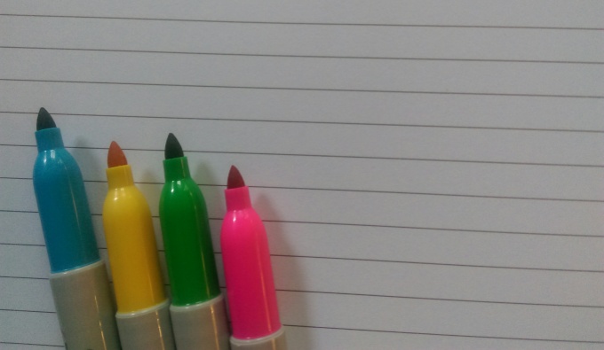 A blue, yellow, green and pink pen in a line. Resting on lined white paper.