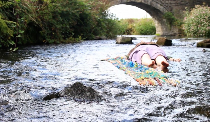 A river with a figure lay down in the water. Toes facing the viewer and head towards three large, stepping stone-like rocks and a large stone bridge in the distance. Next to the figure, stretched out in the water is a piece of loosely woven textile. 