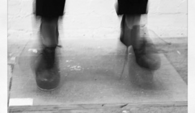 A lack and white image of tap dancing feet on a board. The image is blurred depicting rapid movement.