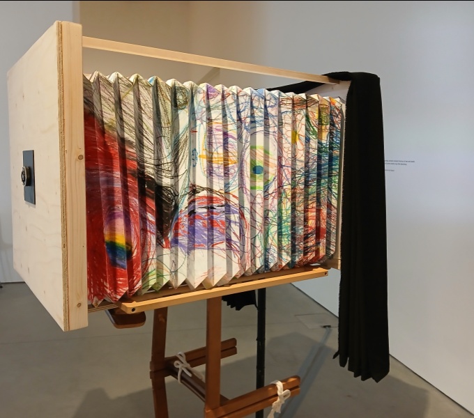 A replica old fashioned camera in enlarged proportions. The corrugated sides made of paper decorated in crayons with multiple colours