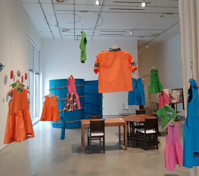 A wide view of the exhibition. Including childrens outfits hanging from the ceiling, a long blue curtain in a spiral, wooden tables and tambourines hanging from the gallery wall.