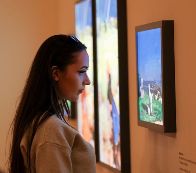 A young woman with a light coloured top and long dark hair looks at artwork on a gallery wall. 