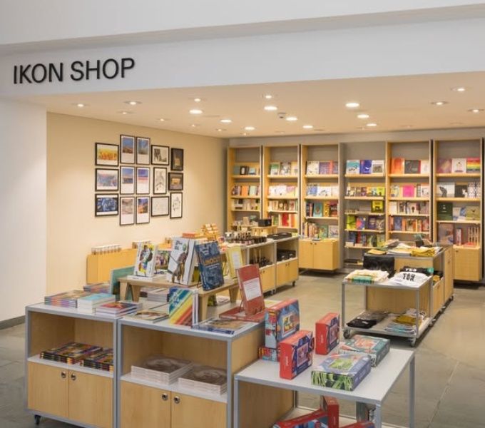 A wide view of the Ikon Gallery shop. It has white walls and ceiling, several wooden shelves laden with books and merchandise, posters displayed on the wall to the left and in the middle are further storage and display units.  