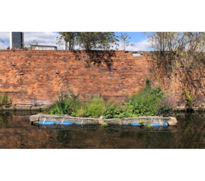 A canal with a red brick wall on the far side. In the centre of the image is a large rectangular shaped vessel filled with wild flowers. 