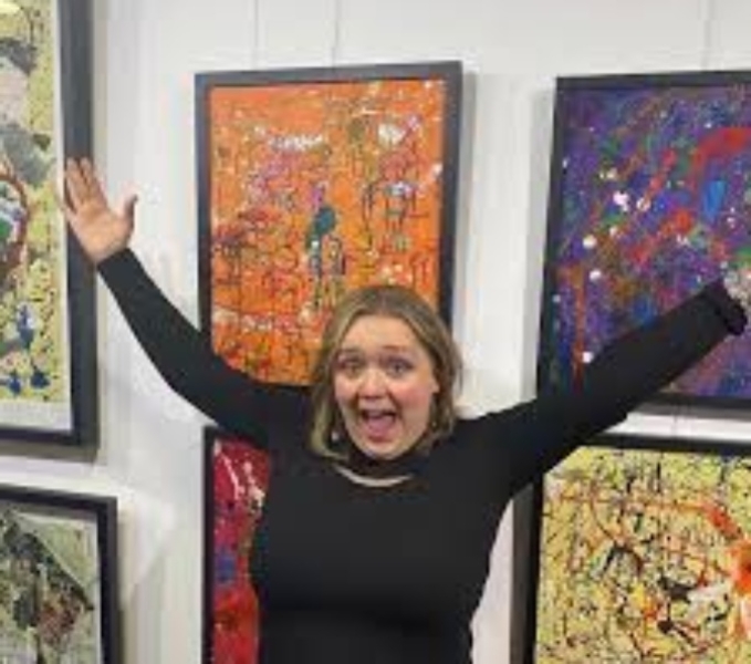Grace Currie wears a long sleeved black top. She has her hands in the air as she looks to the camera. Behind her, several framed artworks are hung on a white wall. 
