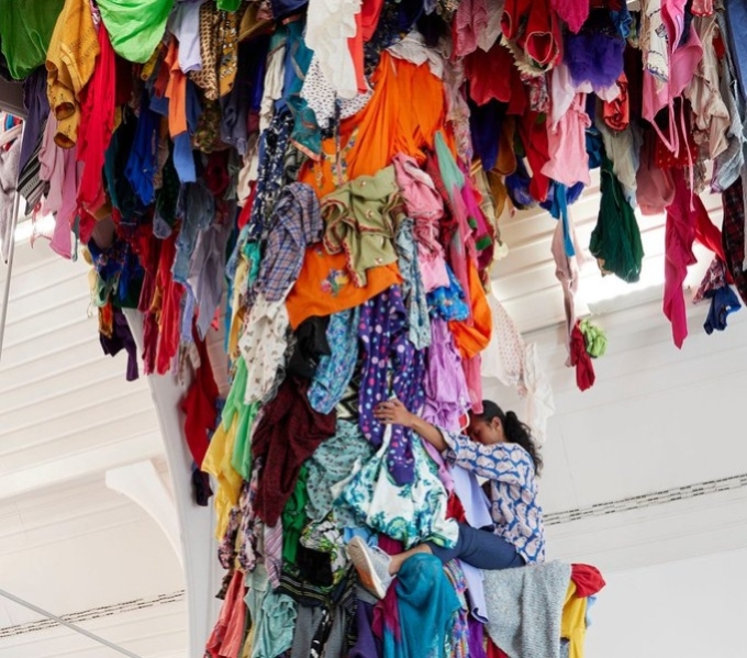 A white gallery space with a tree like form hanging with multicoloured cloth and clothing. Towards the top a young woman wraps her legs around the trunk like structure and holds tight. 