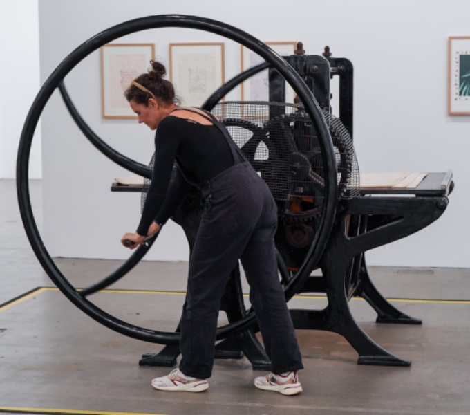 A large black antique press in a gallery space. A lady wearing dungarees turns the large handle.