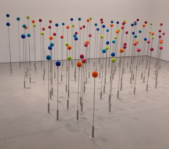 A white gallery space with 100 woollen pom poms arranged on metal poles. The pom poms are neat and tightly clipped in colours orange, pink, green, blue and turquoise.