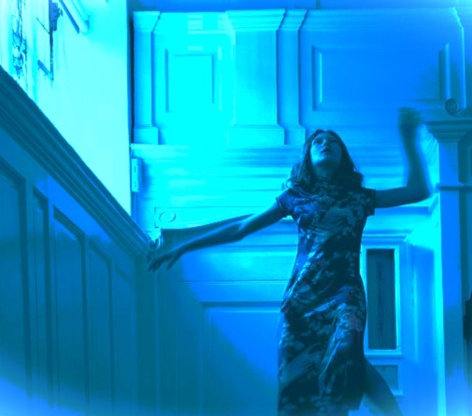 A beautiful old room with white panelled walls and an ornate, curved ceiling. A high window on the left has stained-glass. Light from this falls onto a young woman. She is suspended in mid-air. She looks distressed. Her legs and arms are flailing.