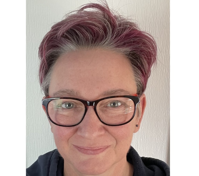 Paula Dower looks up at the camera and is smiling. She wears dark rimmed glasses and has short cropped hair that us dyed a deep pink/ purple colour. She wears a black tshirt and navy hoodie.