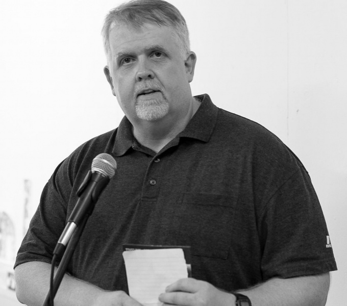 Black and white photographic portrait of Peter Bonnell, from the waist up. Peter has short hair and a short beard, wears a dark polo shirt and stands before a microphone holding a lined notepad between his hands, and wears a digital wristwatch.