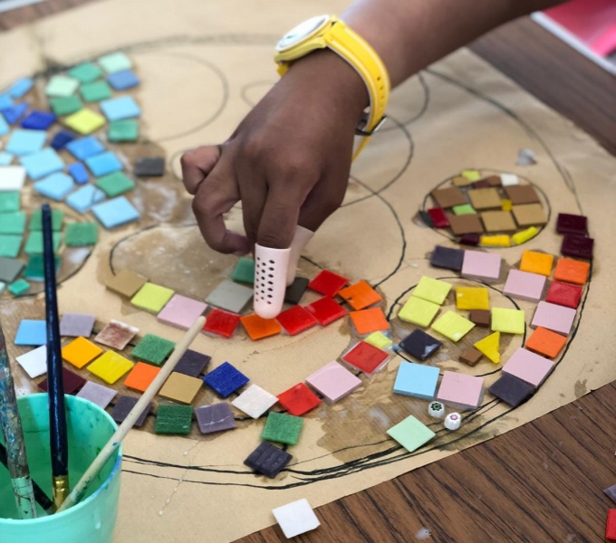 Image shows a close up of the arm of a black person wearing a yellow watch arranging colourful mosaic tiles on a board.