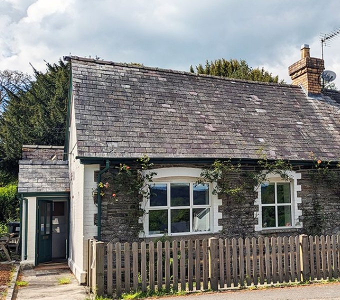 An old grey stone building with slate roof. A picket fence runs along the front of the building. It is a sunny day and she sky is blue but cloudy. 