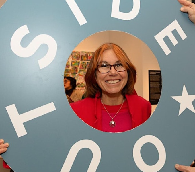 A woan with brown hair, glasses and red top peers through a blown up circular cut out of the Outside In logo.