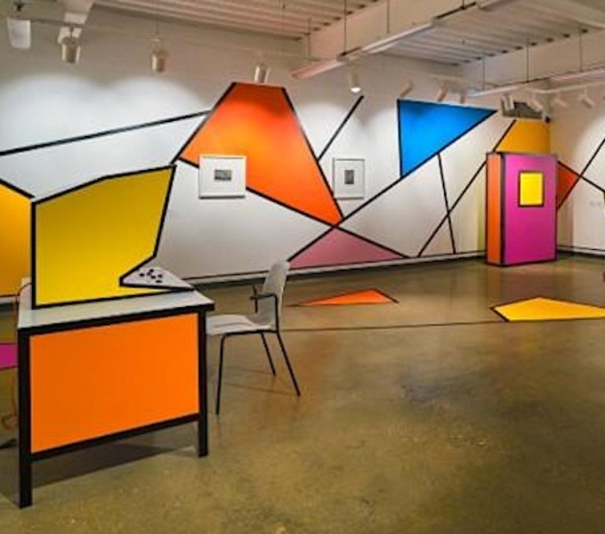 A white gallery space with a wooden floor. The walls and floor are covered in bright geometric shapes in blue, pink, yellow and orange, created within black lines. There is what looks like a retro yellow computer game in the centre of the room.