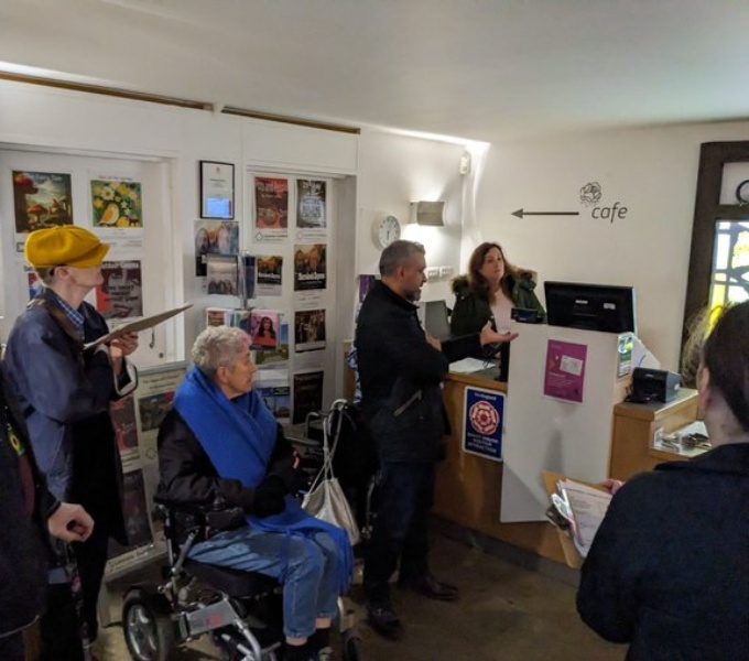 A crowd of people in what looks like the reception area of a tourist venue, museum or gallery. One person is using a wheelchair and the rest are standing as the listen to the person behind the desk. 