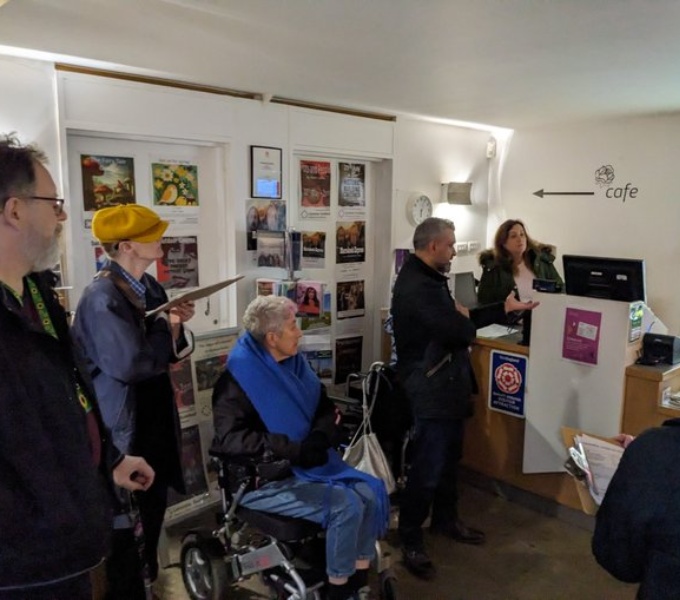 A crowd of people in what looks like the reception area of a tourist venue, museum or gallery. One person is using a wheelchair and the rest are standing as the listen to the person behind the desk. 