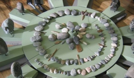 This landscape formatted image shows a circular matt green disc on a wooden floor. On the disc is an arrangements of pebbles in a wide spiral. This surrounds an inner arrangement of pebbles in a starburst formation in the centre.