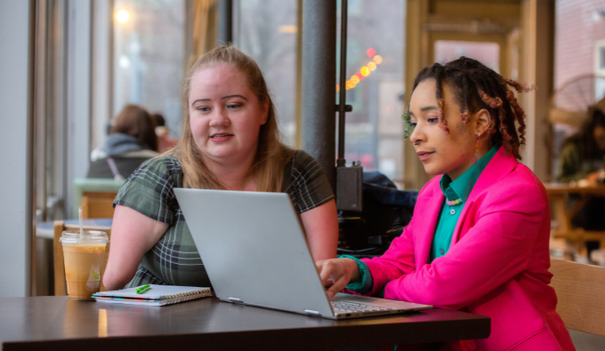 Image shows two people sat at a table using a laptop computer.